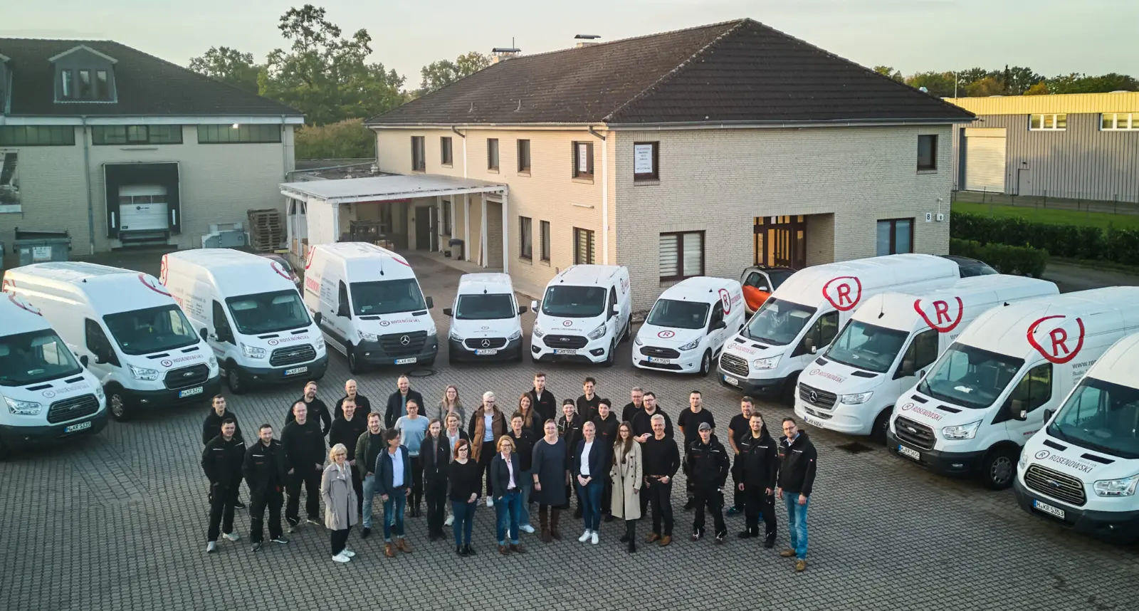 Gruppenbild des Küchen Rosenowski Teams aus der Vogelperspektive mit weißen Transporten und Rosenowski Design im Hintergrund